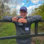 Jacques holds his camera and leans against a tall gate, smiling at the camera and wearing a blue Nova Scotia Nature Trust ball cap.