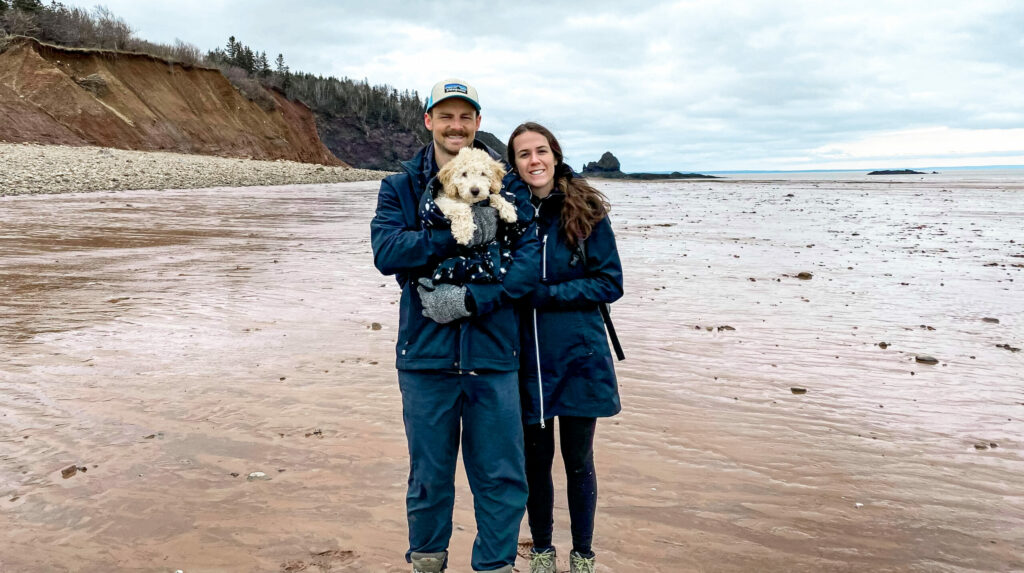 Mark and Hilary standing with their puppy along a sandy beach at low-tide
